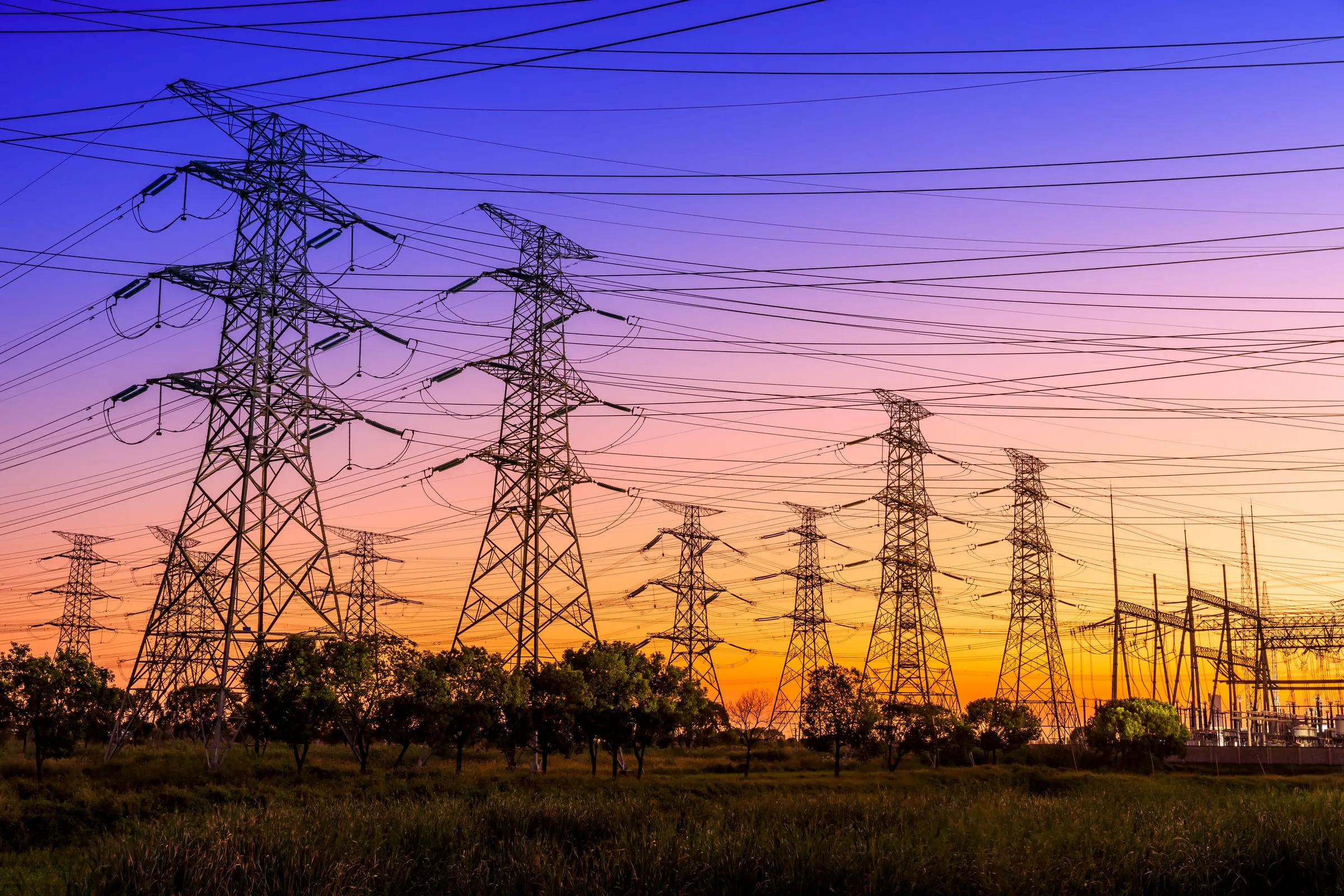 Rows of high-voltage transmission towers silhouetted against a sunset sky that shifts from deep violet overhead to orange at the horizon, their lines receding toward a substation at the right